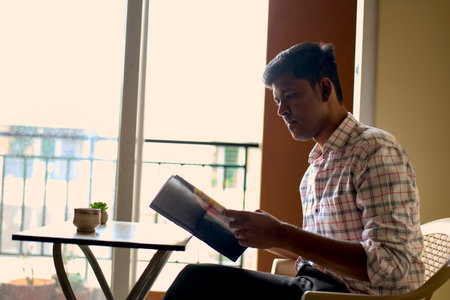 A young man sits quietly at a table, engrossed in a magazine as sunlight filters through a nearby...