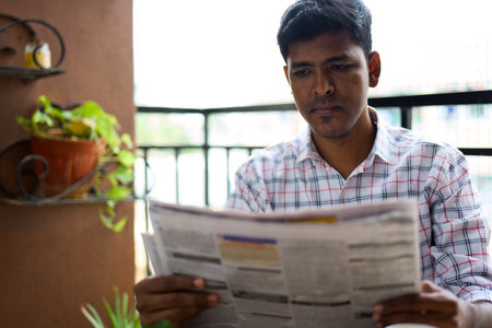 A young man sits on a balcony, engrossed in reading a newspaper. The vibrant greenery around him adds a lively touch to the sunny morning atmosphere.の写真素材