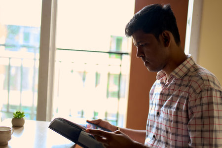 A young man is deeply engaged while reading on a book indoors, illuminated by soft, natural light streaming through a window, highlighting concentration.の写真素材