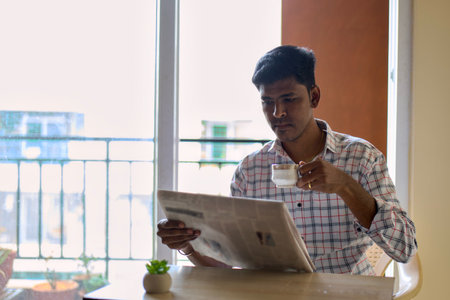 In the morning light, a young man sits at a table, savoring tea while reading a newspaper, immersed...