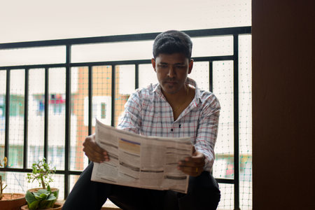 The young man sits comfortably on a balcony, focused intently on reading a newspaper. Potted plants add a touch of greenery to his urban environment during the late afternoon.の写真素材