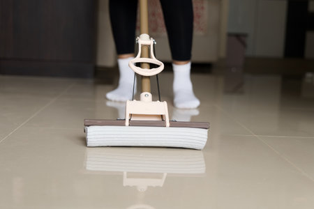 A close-up image of a person using a mop to clean a tiled floor. The focus is on the mop and the...