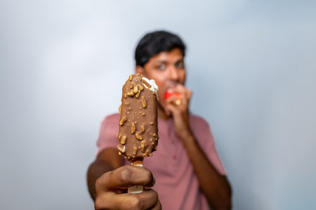 Chocolate-covered ice cream bar with nuts, held out by a young man enjoying a sweet treat. Perfect for summer, dessert, or snack themes.の写真素材
