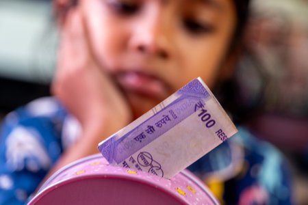 Young child thoughtfully inserting Indian rupees into a pink piggy bank, learning about saving money.の写真素材