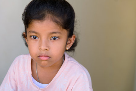 A young girl with dark hair and a serious expression sits quietly against a plain background. She wears a light-colored shirt and looks off to the side, deep in thought.の写真素材