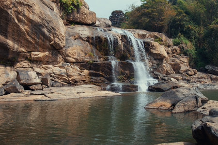 This image captures the tranquil scene of a waterfall cascading down a rocky cliff into a calm pool below.の写真素材
