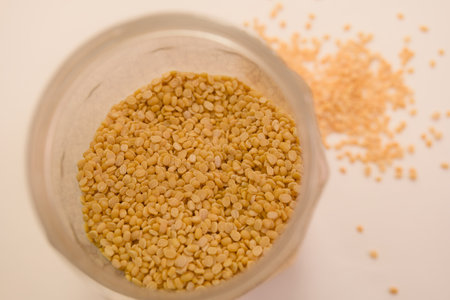 A glass jar holds yellow sesame seeds, with some scattered on a white surface.