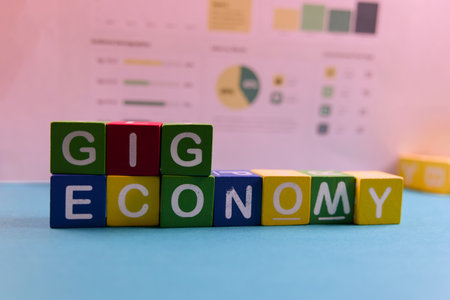 Colorful building blocks spell out gig economy on a desk with charts and graphs in the background, showcasing data.の写真素材