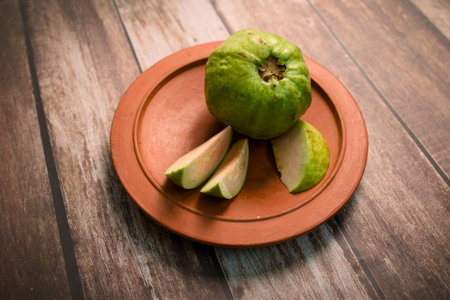 A whole quince fruit sits on a clay plate next to sliced pieces on a wooden table surface.の写真素材