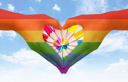 Hands painted as the rainbow flag forming a heart, symbolizing gay love on blue sky. Fredom concept.