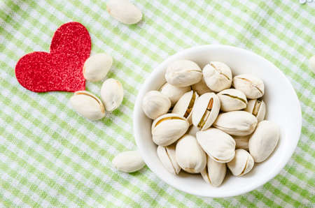 Pistachio nuts in white bowl and red heart shape paper on checkered clothの写真素材