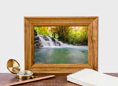Beautiful waterfall in photo frame with vintage compass, diary note and pencil on wooden table. Travel concept.の写真素材