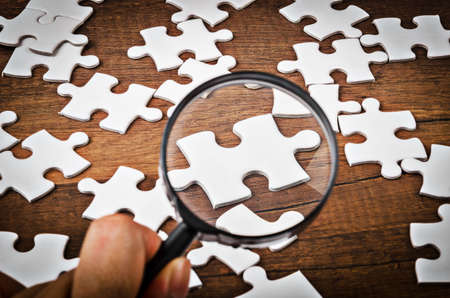 Hand with magnifying glass on white jigsaw puzzle on wooden background.の写真素材