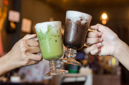 Friends toasting, saying cheers holding glass of iced chocolate and green tea matcha in restaurant.