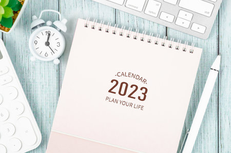 2023 Desk calender with alarm clock, keyboard computer, pen, plant pot, and calculator on blue...