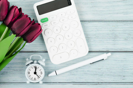 Flat lay, top view office wooden table desk. Workspace with Alarm clock, calculator, pen an red tulip flower on blue wooden background.の写真素材
