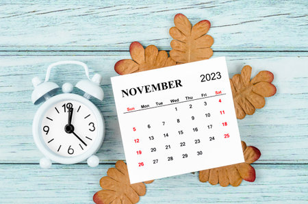 November 2023 Monthly calendar year and alarm clock with dried leaves on blue wooden background.の写真素材