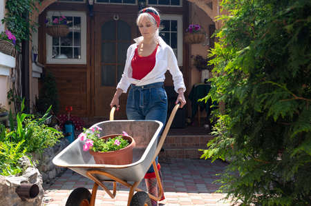 Beautiful blond middle aged woman in white shirt and blue jeans pushing a handcart with flowerの写真素材