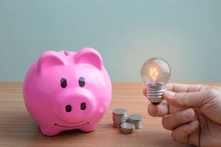A man's hand holding a light bulb with a light background with a pink piggy bank and a coin placed...