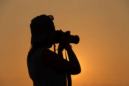 silhouette of a woman holding camera taking pictures outside during sunrise or sunset. Travelling and vacation concept / holiday travelingの写真素材