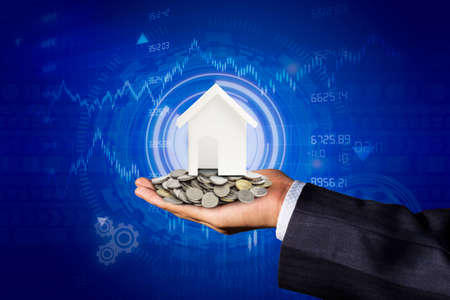 The hand of a businessman with coins on his hands and a model house placed on a coin. Blue background, business concept, real estate land tradingの写真素材