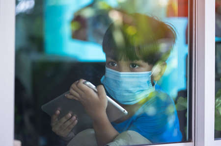 Cute asian young boy with blue shirt, wearing medical mask, sitting near window with tablet....