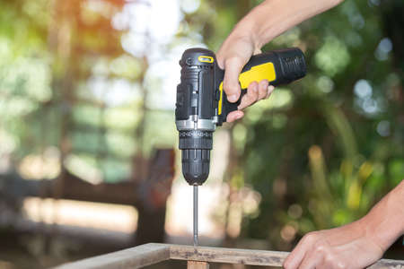 Skilled professional carpenter in workwear using electric drill machine, custom woodworking, making woodware, young experienced craftsman drilling wooden planks on table at workshop. Small businessの写真素材