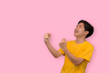 Happy excited young Asian man raising his fists doing yes gesture celebrating success in empty space aside in isolated studio pink backgroundの写真素材