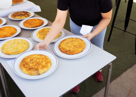 This image features a person serving multiple plates of traditional Spanish omelette (tortilla espanola) at an outdoor event. The scene captures a variety of omelettes arranged on a table, ready to be served. The person is holding a plate and a napkin, demonstrating the communal and inviting nature of the event. The assortment of omelettes showcases different variations of this classic dish, making this image perfect for illustrating traditional Spanish cuisine and communal diningの写真素材