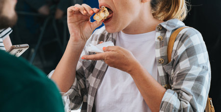 This close-up image captures a woman enjoying a pincho of Spanish omelette (tortilla espanola). She is holding the omelette with her fingers and about to take a bite, demonstrating a casual and enjoyable dining moment. The woman is dressed in a plaid shirt over a white t-shirt and holds a napkin in her other hand. This image highlights the cultural tradition of eating pinchos in Spain and the joy of sharing simple, delicious foodの写真素材