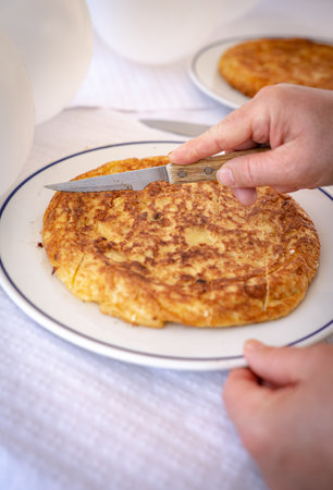 This image shows a close-up of hands slicing a traditional Spanish omelette (tortilla espanola) with a wooden-handled knife. The omelette is placed on a white plate with a blue rim, and white balloons are visible in the background, indicating a festive or celebratory event. The golden-brown color of the omelette and the festive decor create a warm and inviting atmosphere. This image is perfect for highlighting traditional Spanish cuisine in a celebratory contextの写真素材