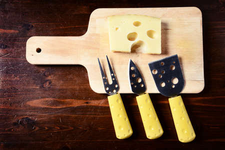 small cheese knives  and chopping board with emmenthal cheese on dark wooden tableの写真素材
