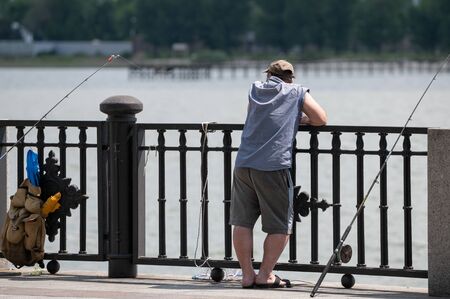 a male fisherman in a cap on the embankment of the river with a fence stands near fishing rods and a...