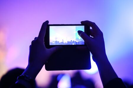 hands from the crowd take videos and photos on a smartphone as a keepsake of a music festival. blurred background. atmosphere of a musical concertの写真素材