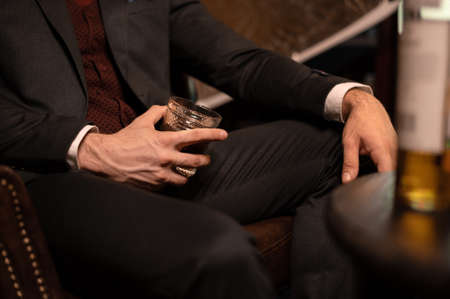A rich man in an expensive suit sits in a dark room with a glass of whiskey. Alcohol in a glass close upの写真素材