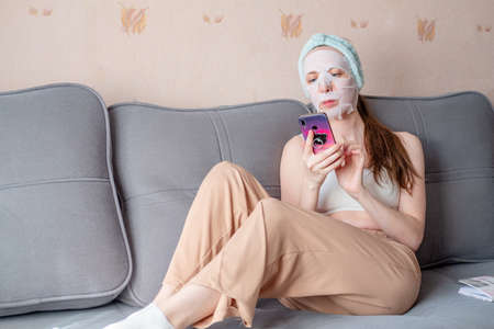 girl in a cosmetic mask lies on the couch and looks at the phone. home treatments for the face. freelancer at work. tkneya msk for the face. work from homeの写真素材