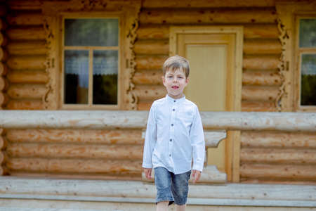 boy walks near the house. Child on stage. boy on the background of a wooden house. Boy in white shirtの写真素材