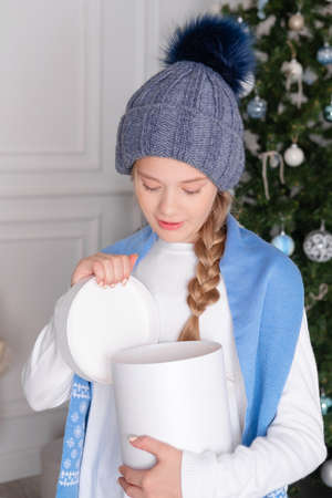 girl opens a gift box. Surprised by the gift. A Christmas gift in the hands of a girl. Surprise for the child. Christmas clothes and interior. Winter hat with pompomの写真素材