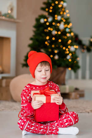The boy opens a Christmas present. Boy in Christmas pajamas with a gift. Fireplace and Christmas tree. Christmas decorations at home. morning unpacking giftsの写真素材