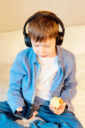 A teenage boy in jeans sits on the bed and listens to music with wireless headphones. Free time....