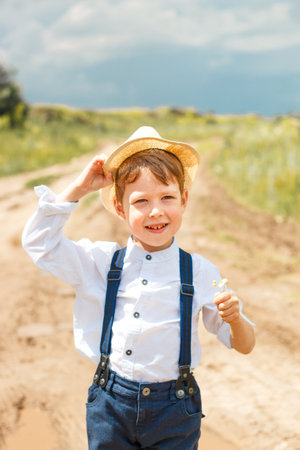 Little farmer on a summer field, cute little boy in a straw hat. boy with a flower stands in a field. boy in rubber boots and a white shirt. Country styleの写真素材
