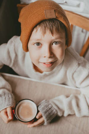 a boy in a sweater and a brown hat drinks cocoa from a red cup. A cozy photo with a mug in hand. boy...