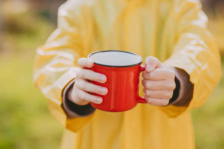 the hands of a boy in a yellow raincoat are holding hot cocoa in a red mug. Cozy photo with a mug. camping concept. Autumn photo with a cup of cocoa on the streetの写真素材