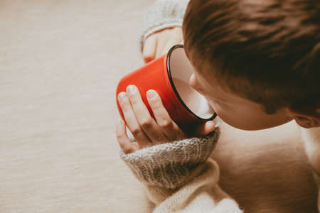 hands in a sweater hold hot cocoa, in a red mug, top view. A cozy photo with a mug in hand with copy space. Child drinks hot chocolateの写真素材