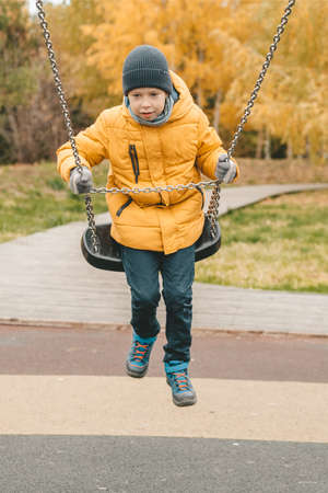 happy boy in a hat and a yellow jacket, riding on a swing, On the street. Walk in the street in...