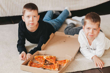 two boys lie on the floor eating pepperoni pizza from a box. vertical frame. two boys lie on the floor eating pepperoni pizza from a box.の写真素材