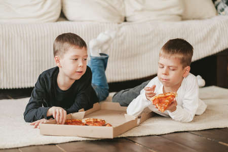 two boys lie on the floor eating pepperoni pizza from a box. vertical frame. two boys lie on the...