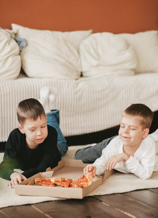 two boys lie on the floor eating pepperoni pizza from a box. vertical frame. two boys lie on the floor eating pepperoni pizza from a box. vertical frameの写真素材
