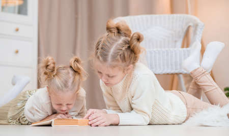 2 blonde girls lie on the floor by the Christmas tree and read a book. cozy evenings with a book by...