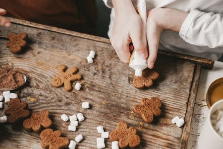 Children decorate the Christmas gingerbread man with white frosting on a wooden background. Christmas sweets with cocoa and marshmallows for children. Winter vacation at home. Fun time in the kitchen.の写真素材